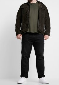 Uomo che indossa una giacca di velluto a coste marrone scuro, una maglietta verde oliva, jeans neri con orli arrotolati e sneakers bianche, in posa davanti a uno sfondo semplice.