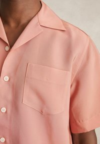 Kortärmad button-up skjorta i mjuk rosa tyg, med en spetsig krage och en enkel bröstficka med subtila linjedetaljer.