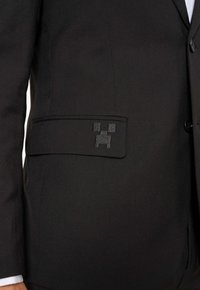 Čierna sako so zreteľnou vreckom, ktoré má pixelovaný čierny dizajn Creepera. Hladký textil, strihaný strih a štruktúrovaná forma.