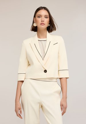 Kobieta w kremowej skróconej marynarce z czarną lamówką, dopasowanych spodniach z wysokim stanem, pod spodem w paski, oraz dużych okrągłych kolczykach.