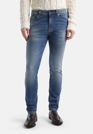 Slim-fit, lichtblauwe denim jeans met een vervaagde wassing, voorzien van vijf zakken, riemlussen en een knoopsluiting. Draagt bij bruin lederen schoenen.