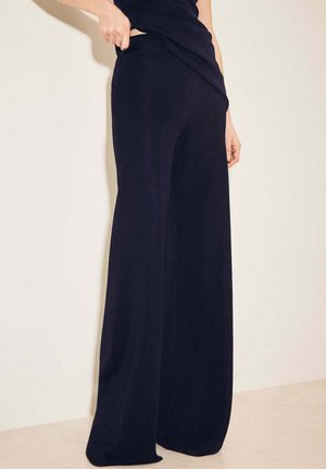 Osoba nosząca czarne spodnie o szerokich nogawkach, ciągnąca za pas, aby pokazać dopasowanie, stoi na tle jasnej ściany i podłogi.