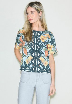 Pitkähiuksinen nainen, jolla on päällään lyhythihainen kukkakuvioinen paita ja vaaleansiniset farkut, seisoo yksiväristä valkoista taustaa vasten.