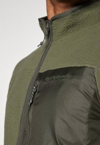 Giacca in pile verde oliva con pannello in nylon liscio, tasca con zip, tessuto strutturato e logo "PeakPerformance" lievemente impresso.