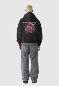 Svart huvtröja med grafisk design på baksidan, med texten "YOUTHQUAKE" och abstrakta element i rosa. Kombinerad med grå jeans.