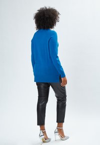 Maglione blu a maglia con collo alto, orlo a coste e spacchi laterali, abbinato a pantaloni di pelle neri e scarpe con tacco alto in argento.