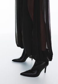 Pantofi cu toc înalt din piele neagră, cu vârf ascuțit, asortați cu o ținută dintr-un material negru translucid, care evidențiază o textură fluidă și un design stratificat.