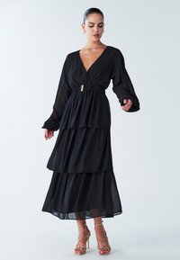 Schwarzes, mehrlagiges Maxikleid mit langen Ärmeln, tiefem V-Ausschnitt, elastischem Taillenbund und goldfarbenem Gürtelakzent. Fließender, leichter Stoff.