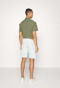 Chemise verte à manches courtes, short en denim bleu clair et baskets beiges. Vue de dos montrant le col de la chemise et l'ourlet court, shorts avec poches.