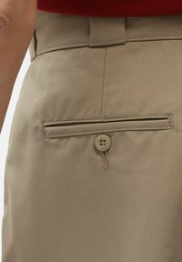Beige trousers featuring a back pocket with a button closure. The fabric has a smooth texture with subtle diagonal patterns.