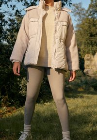 Dikke crèmekleurige jas met een textuur van beige fleece aan de bovenkant, een volledige rits en vier zakken; gecombineerd met lichtbeige geribbelde leggings.