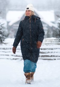 Dikke marineblauwe winterjas met een rits, bruine accenten, blauwe jeans en tan kleurige laarzen. Een witte beanie maakt de outfit compleet in een besneeuwde omgeving.