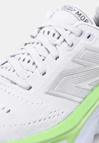 White sneaker with mesh and synthetic upper, reflective silver accents, and a green EVA midsole. Features a lace-up design and perforated details.