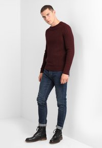 Burgunderfarget strikket genser med teksturert mønster, kombinert med mørke jeans og svarte snøreboots. Genseren har rund hals og ribbestrikkede mansjetter.