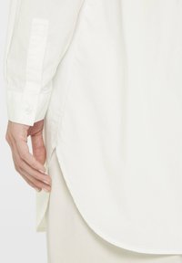 White long-sleeve shirt with buttoned cuff, featuring a curved hem and soft fabric texture. A hand touches the side seam, showing detail.