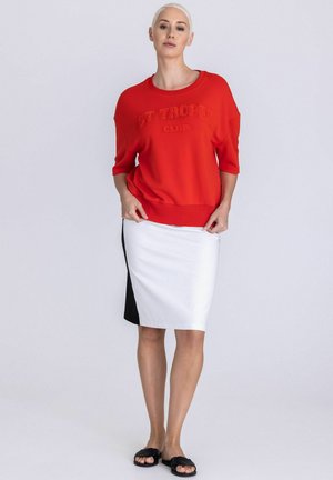 Femme aux cheveux blonds courts portant un haut rouge "St. Tropez Club", une jupe blanche avec un panneau latéral noir et des sandales noires à bout ouvert, debout devant un fond uni.