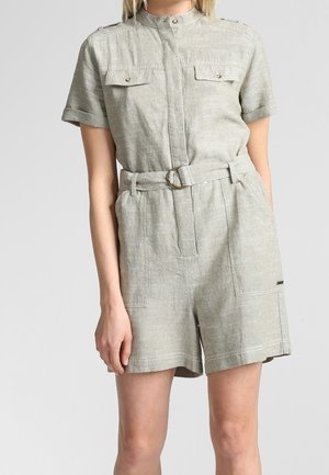 Mujer vestida con un mono corto de lino gris claro con manga corta, cinturón, bolsillos en el pecho con botones y cuello mao.