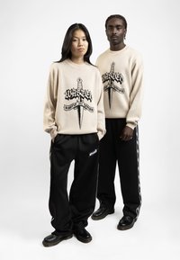 Beige trui met zwart grafisch ontwerp waarop een dolk en tekst staan; gecombineerd met zwarte broek met zijstrepen en zwarte schoenen.