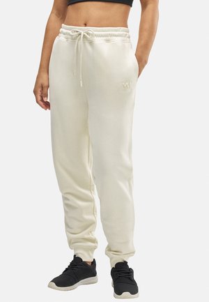 Pantalones de chándal color crema con cintura ajustable, puños acanalados y un logo sutil en el muslo, combinados con zapatillas deportivas negras.