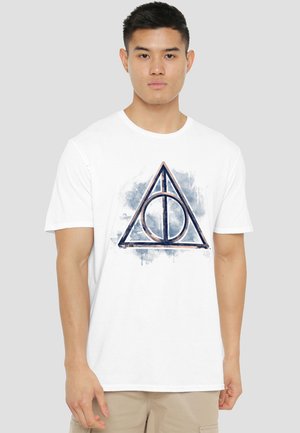 Young man wearing white T-shirt with a large blue and gray triangular symbol featuring a circle and vertical line within.