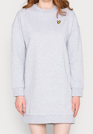 Kobieta ubrana w jasnoszarą sukienkę-bluzę z długim rękawem, z małym żółtym logo ptaka na górnej lewej części klatki piersiowej.