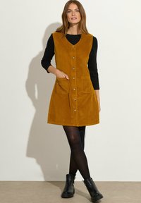 Vestido de pana mostaza con escote en V, cierre frontal de botones y dos bolsillos de parche. La modelo lleva una camiseta de manga larga negra y medias, combinadas con botas.