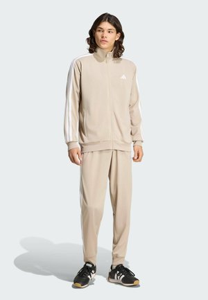 Νέος άνδρας στέκεται, φοράει μπεζ φόρμα Adidas με λευκές ρίγες και μαύρα αθλητικά παπούτσια Adidas, κοιτάζοντας ελαφρώς προς τα πλάγια.