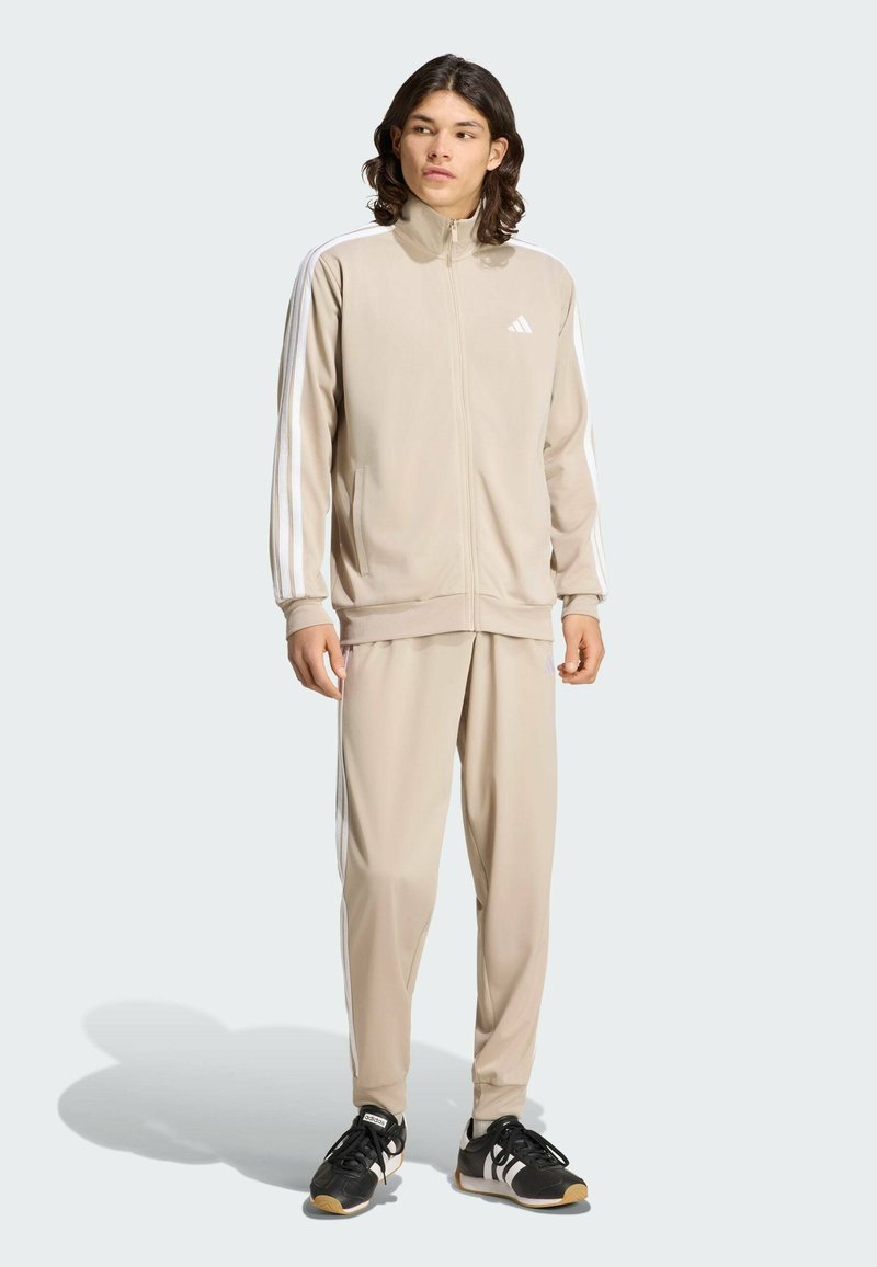 Νέος άνδρας στέκεται, φοράει μπεζ φόρμα Adidas με λευκές ρίγες και μαύρα αθλητικά παπούτσια Adidas, κοιτάζοντας ελαφρώς προς τα πλάγια.