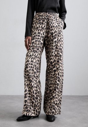 Vrouw draagt wijde broek met luipaardprint, zwarte top met lange mouwen en zwarte schoenen, staand op een grijze vloer tegen een grijze muur.