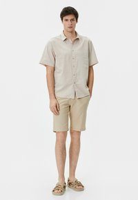 Korte mouwen, beige knoopshirt met een gestructureerde afwerking, gecombineerd met beige shorts, gedragen met tan sandalen. Eenvoudig, casual ontwerp.