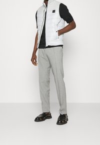 Vadderad vit väst med hög krage, dragkedja och logodetalj, i kombination med grå byxor och svarta snörskor. Enkel, modern design.