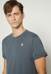 Tricou gri din bumbac, cu decolteu rotund și mâneci răsucite, prezintă un mic logo alb în zona stângă a pieptului. Textură netedă.