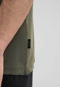 Olivgrünes kurzärmliges Hemd aus strukturiertem Material, mit einem schwarzen Etikett an der Seitennaht und sauber genähten Säumen.