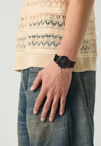 Montre chronographe noire au poignet, avec un cadran rond, un bracelet en maille noire et un cadran détaillé comportant trois sous-cadrans. Portée avec un haut en maille.