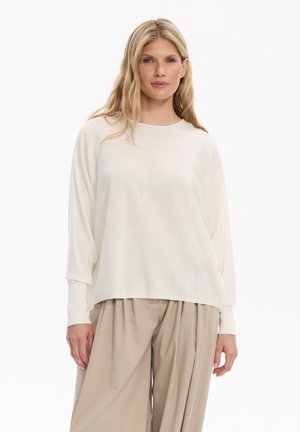 Kobieta z długimi blond włosami, ubrana w luźną białą bluzkę z długim rękawem i beżowe szerokie plisowane spodnie na jednolitym białym tle.