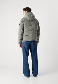 Puffjakke i lys grå stof med hætte, med vertikal ribbet tekstur og et mærke på venstre ærme, parret med vide blå jeans.