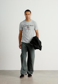 T-shirt cinza de algodão com o logótipo "Gant 1949", combinada com jeans azul folgado, jaqueta preta na mão e sapatos escuros com solas claras.
