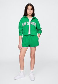 Felpa verde corta con cappuccio con scritta "GAP SPORT" in rosa, abbinata a pantaloncini verdi con rifiniture rosa e sneakers bianche.