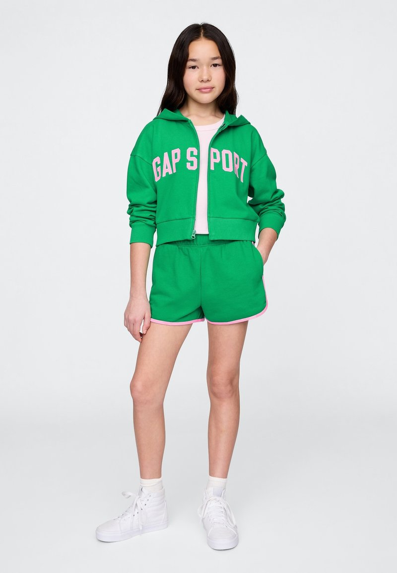 Felpa verde corta con cappuccio con scritta "GAP SPORT" in rosa, abbinata a pantaloncini verdi con rifiniture rosa e sneakers bianche.