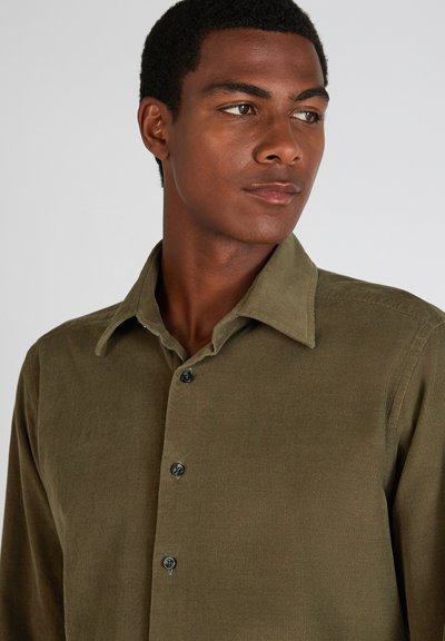 Giovane uomo con capelli corti e ricci che indossa una camicia a sbottonatura in velluto a coste verde oliva, che guarda di lato su uno sfondo semplice.