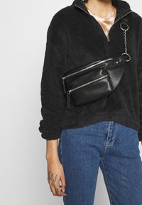 Sudadera de forro polar negra con collar con cremallera, combinada con una riñonera de cuero negro que cuenta con varios bolsillos con cremallera y un detalle de cadena de metal.