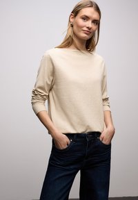 Jeune femme aux cheveux blonds portant un pull beige à manches longues et un jean bleu foncé, mains dans les poches, debout devant un fond uni.
