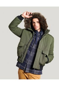 Oliven grøn pufferjakke med en faux fur-fôret hætte, der har lynlåslukning, to frontlommer og en flannel skjorte under.