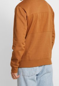 Muž oblečený v hnědé mikině s dlouhým rukávem a světle modrých džínech, zobrazený zezadu na bílém pozadí.