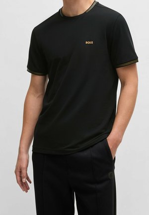 Mężczyzna ubrany w czarny krótki rękaw T-shirt z logo "BOSS" na piersi i czarne spodnie, stoi z jedną ręką w kieszeni na jednolitym tle.
