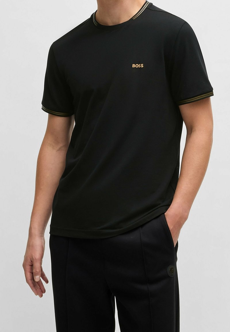 Homme portant un t-shirt noir à manches courtes avec le logo "BOSS" sur la poitrine et un pantalon noir, debout avec une main dans la poche sur un fond uni.