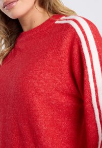 Pull en tricot rouge avec un col rond, présentant deux rayures blanches le long des épaules. Texture douce et coupe décontractée visibles.