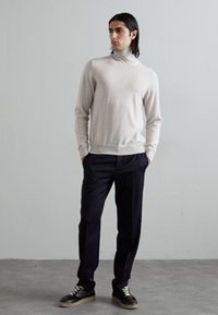 Maglione a collo alto grigio chiaro, design aderente, abbinato a pantaloni scuri e sneakers nere con suola bianca, in piedi su uno sfondo neutro.