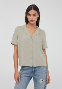 Blusa grigia chiara a maniche corte con colletto a punta, chiusura frontale con bottoni e vestibilità comoda, abbinata a jeans azzurri distressed.