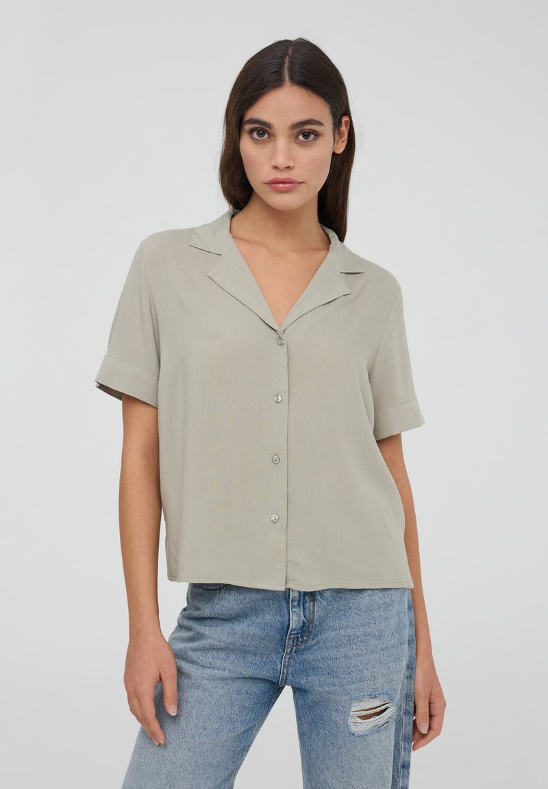 Blusa grigia chiara a maniche corte con colletto a punta, chiusura frontale con bottoni e vestibilità comoda, abbinata a jeans azzurri distressed.